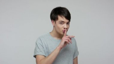 Shh, be quiet, don't tell secret! Positive brunette man in T-shirt pointing me and you, showing silence gesture and asking keep silent about our affair. indoor studio shot isolated on gray background