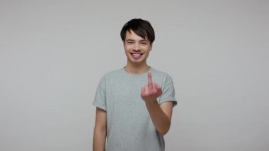 Crazy careless cheerful man in T-shirt showing middle fingers to everyone around, gesturing fuck you all and smiling, expressing indifference to problem. indoor studio shot isolated on gray background