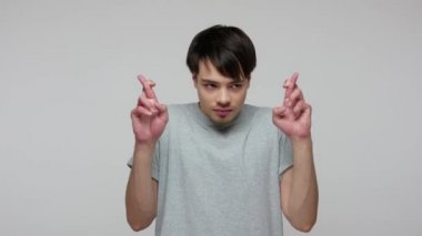 Unhappy young brunette man in T-shirt praying before important occasion and holding fingers crossed for good luck, hoping for victory, wishing miracle. indoor studio shot isolated on gray background
