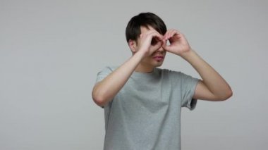 Young man holding hands in binoculars gesture, observing area around with serious attentive look, expressing shock and surprise when focuses on camera. indoor studio shot isolated on gray background