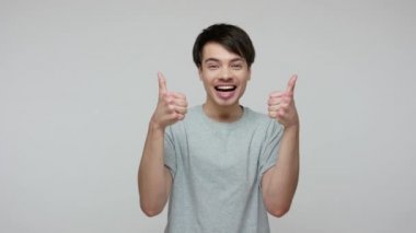 Excited young brunette man in T-shirt shouting Yeah, enjoying success and showing thumbs up, double like gesture, satisfied with awesome service. indoor studio shot isolated on gray background