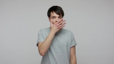 Shocked frightened young man in T-shirt covering mouth with hand and looking terrified at camera, scared to tell truth, intimidated bullied victim. indoor studio shot isolated on gray background