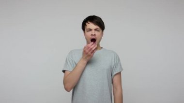 Young exhausted student guy in T-shirt yawning and stretching hands up, feeling lazy drowsy, tired after sleepless night of study, suffering insomnia. indoor studio shot isolated on gray background
