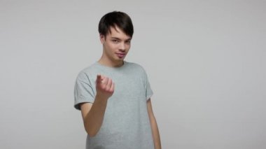 Hey beauty, come here! Handsome brunette man in T-shirt flirting and gesturing with one finger, making beckoning sign, inviting for date and looking with alluring eyes. studio shot, gray background