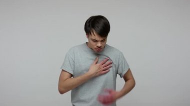 Young guy in T-shirt grabbing his chest and grimacing from sudden pain, symptoms of heart attack, infarction, having lung problems, risk of pneumonia. indoor studio shot isolated on gray background