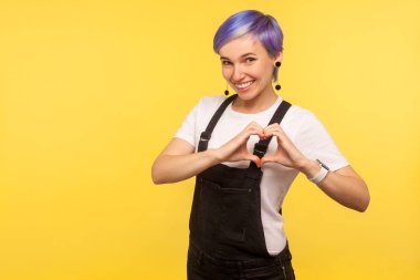 Güzel romantik hippi kız portresi kot tulumlu kısa saçlı, dost canlısı gülümseyen ve kalp sembolünü elleriyle gösteren, aşk işareti olan. İçerde, sarı arka planda izole edilmiş, stüdyo çekimi