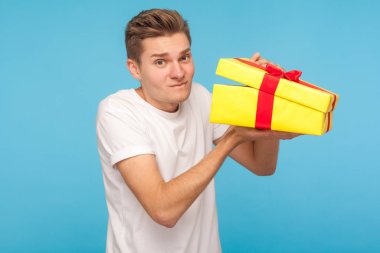 Sıradan beyaz tişörtlü mutsuz bir adam paketlenmemiş bir hediye kutusu tutuyor ve tatminsiz bir ifadeyle kameraya bakıyor, kötü hediyelerden memnun değil. Mavi arka planda kapalı stüdyo çekimleri