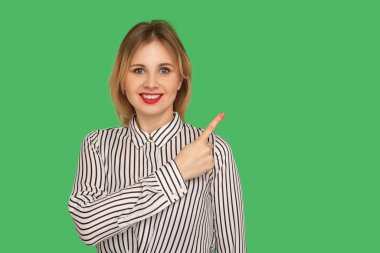 Çizgili bluzlu, kırmızı dudaklı harika bir kız boş fotokopi alanını işaret ediyor ve samimi bir şekilde gülümsüyor, reklam içeriği için yan boş duvarı gösteriyor. Yeşil arka planda izole edilmiş kapalı stüdyo çekimi