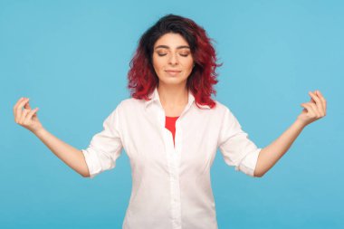 Relaxation and harmony. Portrait of peaceful hipster woman with fancy red hair keeping hands in mudra gesture, practicing breath techniques, meditation. indoor studio shot isolated on blue background clipart