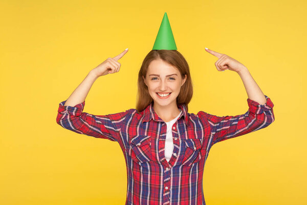 Look at my hat. Portrait of glad excited ginger girl pointing at funny party cone on her head and smiling at camera, celebrating holiday, birthday. indoor studio shot isolated on yellow background