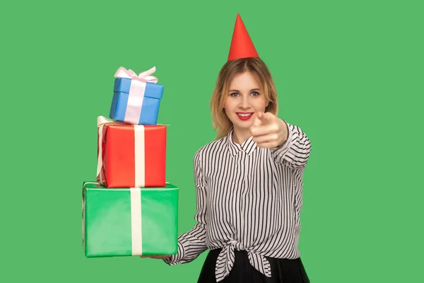 善良的快乐女人 头上挂着滑稽的派对圆锥 指着相机 拿着许多礼品盒 选择你来收到礼物和奖金 祝贺你的生日 工作室拍摄绿色背景 — 图库照片