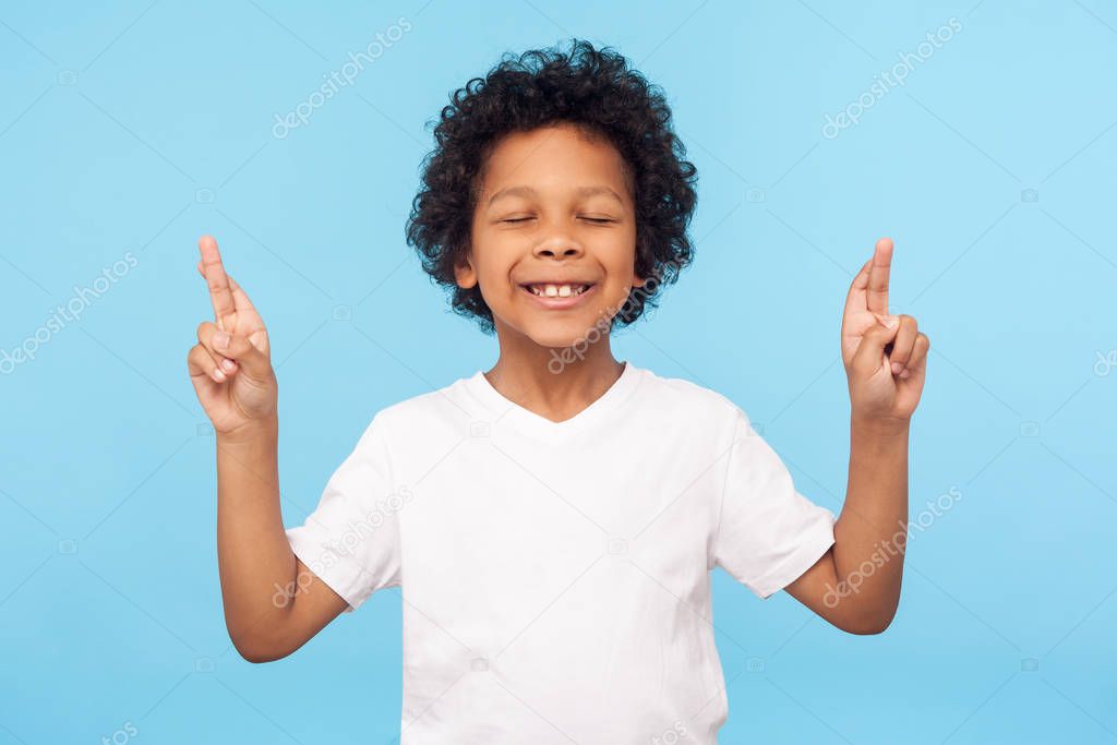 Childhood dreams and hopes. Portrait of adorable little boy standing with closed eyes and crossing fingers, wishing for good luck, waiting miracle. indoor studio shot isolated on blue background