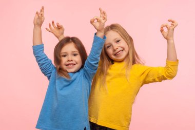 Çocukların durumu iyi, her şey yolunda. İki sevimli mutlu küçük kız parmaklarıyla hareket ediyor ve birlikte gülümsüyor, yani tamam, aynı fikirdeyiz. Pembe arka planda kapalı stüdyo çekimleri
