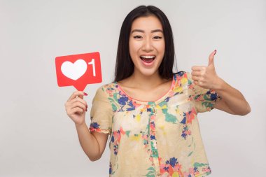 Sosyal medya içeriğine ekle! Neşeli hevesli Asyalı kız internet kalbi ikonu gösteriyor ve başparmağını kaldırıyor, abone olmayı ve ilginç blogu takip etmeyi öneriyor. Kapalı stüdyo çekimi, izole.