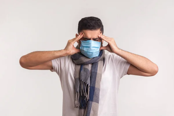 Unhealthy man in surgical mask and scarf suffering headache, rubbing painful temples, having symptoms of flu, contagious coronavirus infection 2019-nCoV. studio shot isolated on white background