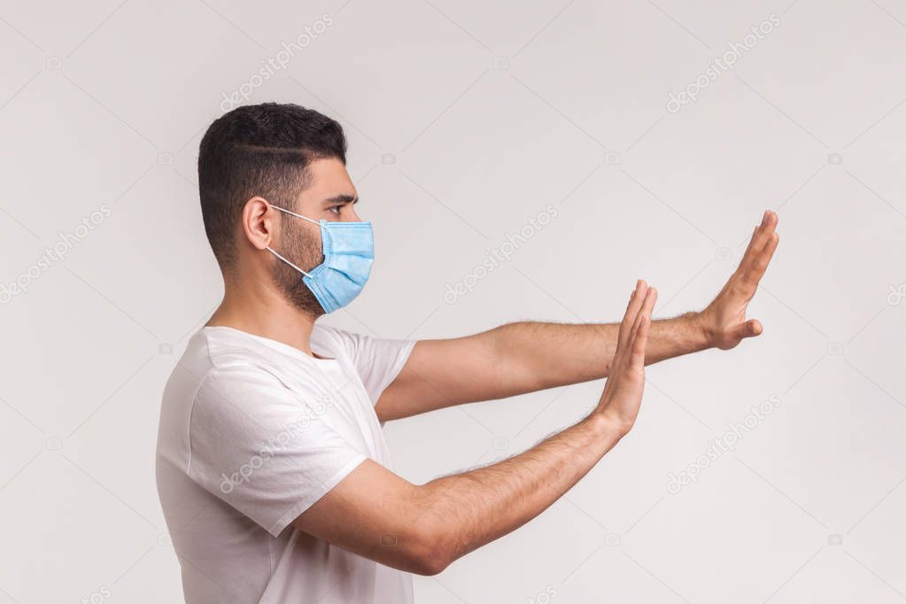 Side view of alarming man in hygienic protective mask gesturing stop, afraid of coronavirus infection, respiratory illnesses such as flu, 2019-nCoV. indoor studio shot isolated on white background