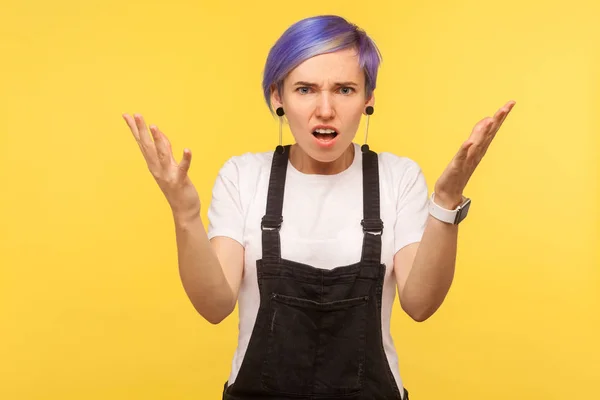 What do you want? Portrait of upset annoyed hipster girl with violet short hair in denim overalls raising hands asking why with indignant face, quarrel conflict concept. yellow background, studio shot