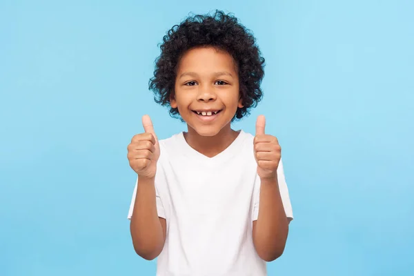 一个快乐的小男孩 卷曲的头发 穿着白色的T恤 对着相机笑着 做着竖起大拇指的姿势 表现出同意酷的认可标志 室内拍摄 蓝色背景隔离 — 图库照片