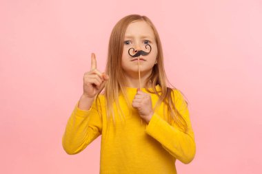 Komik kağıt bıyıklı, patronluk taslayan, jest yapan, tavsiye veren, akıllı erkek gibi davranan maskeli aksesuarla eğlenen küçük kızıl kız. Pembe arka planda kapalı stüdyo çekimleri