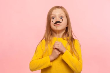 Komik kağıt bıyıklı şirin kızıl bir kızın portresi. Şaşırmış gözlerle kameraya bakıyor, eğleniyor, maskeli aksesuar takıyor. Pembe arka planda kapalı stüdyo çekimleri