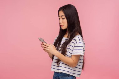Siyah saçlı, tişörtlü, cep telefonuna mesaj yazan sosyal ağda yazı okuyan, ciddi bir ifadeyle internette sohbet eden bir kızın yan görüntüsü. Pembe arka planda kapalı stüdyo çekimleri
