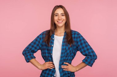Kareli tişörtlü iyi huylu bir kızın portresi kameraya samimi bir şekilde gülümsüyor, kalçalarının üstünde el ele tutuşuyor, kendinden emin ve hayattan memnun görünüyor, pozitif haberler veriyor. Stüdyoda çekim izole edildi