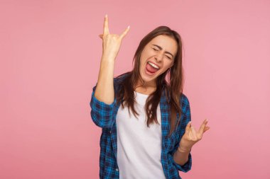 Kareli tişörtlü, coşkulu kızın portresi rock 'n roll jesti gösteriyor ve mutlulukla bağırıyor, festivalde çılgın bir rockçı gibi hissediyor. Stüdyo çekimi pembe arkaplanda izole edildi