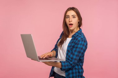 Şaşırmış serbest çalışan portresi, sıradan tişörtlü şaşkın bir kız elinde dizüstü bilgisayar, eğitim veya internet işi için bilgisayar kullanıyor, internette geziniyor. Stüdyo çekimi pembe arkaplanda izole edildi