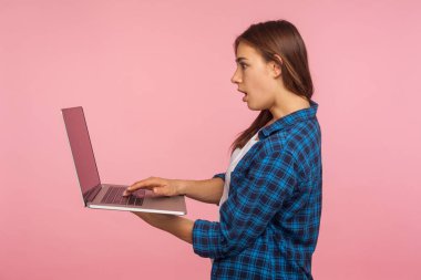 Gelişigüzel tişörtlü, laptoptan e-posta okuyan, hayret verici ifadeleri olan, eğitim için bilgisayar kullanan ya da internet üzerinden iş arayan şaşırmış bir kızın yan görüntüsü. Kapalı stüdyo çekimi, izole.