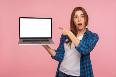 Şok olmuş ifadesiyle boş dizüstü bilgisayar ekranı gösteren kareli tişörtlü büyüleyici kızın portresi, internet reklamcılığı için kopya alanı. Stüdyo çekimi pembe arkaplanda izole edildi