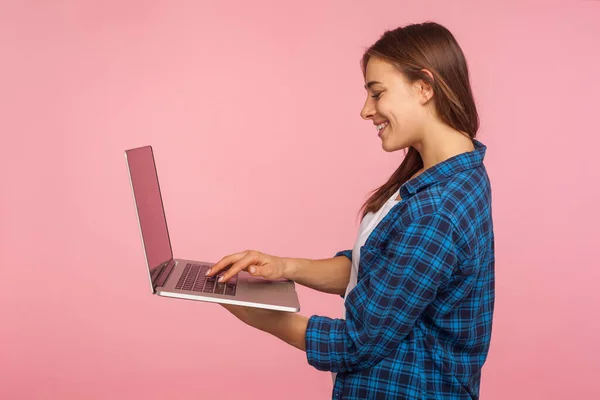 教育やオンラインの仕事のためのコンピュータを使用して ラップトップ上でフリーランスのタスクを行うカジュアルチェックシャツの笑顔の女の子の側面図 インターネットを閲覧 ピンクの背景に隔離された屋内スタジオで — ストック写真