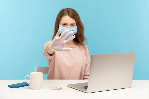 Girl in facial mask and hygienic gloves sitting at desk with laptop, showing stop, warning about danger of getting sick at office workplace, infectious disease, coronavirus epidemic. studio shot