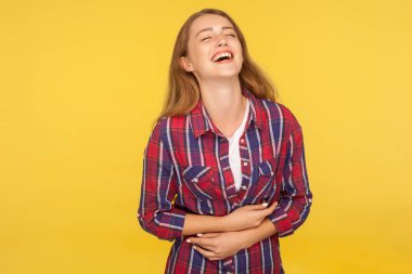 Portrait of carefree ginger girl in checkered shirt holding belly and laughing out loud after hearing hilarious joke, funny anecdote, being in good mood. studio shot isolated on yellow background clipart