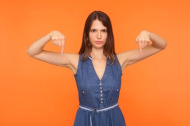 Hemen şimdi ve burada olmalı. Kot elbiseli kızgın esmer kadın iki eliyle aşağıya bakıyor, kontrol etmeye çalışıyor, derhal gelmeyi talep ediyor. Turuncu arka planda kapalı stüdyo çekimleri