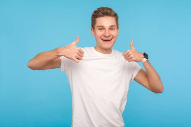 Aferin sana! Mutlu, hevesli, beyaz tişörtlü bir adamın portresi. Başparmaklarını kaldırıyor ve gülümsüyor. Sonuçtan memnun. Mavi arka planda kapalı stüdyo çekimleri