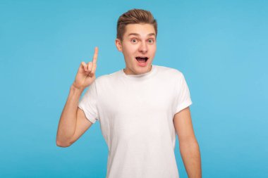 Mükemmel bir fikrim var! Sıradan beyaz tişörtlü heyecanlı yaratıcı adamın portresi parmak kaldırıyor ve aniden icat edilen çözüme şaşırmış gibi bakıyor. Mavi arka planda kapalı stüdyo çekimleri