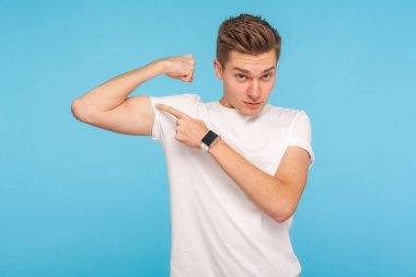 Gücüme bak! Beyaz tişörtlü sıska genç bir adamın portresi. Kollarını kaldırmış, güçlü ve kendinden emin bir şekilde kaslarını gösteriyor. Mavi arka planda kapalı stüdyo çekimleri