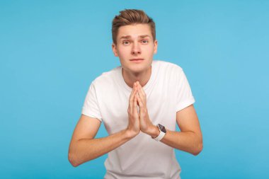 Lütfen, bana bir iyilik yap! Dua ederken el ele tutuşan ve kederli yalvaran bembeyaz tişörtlü yalvaran adamın portresi. Mavi arka planda kapalı stüdyo çekimleri