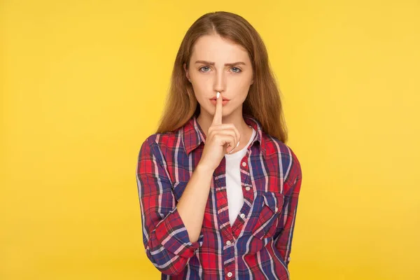 Shh, be quiet! Portrait of serious ginger girl in checkered shirt holding finger on lips making hush silence gesture, asking to keep secret, don\'t speak. studio shot isolated on yellow background