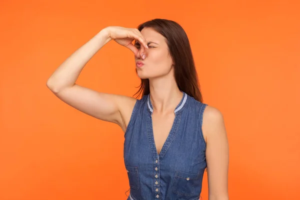 Stink, awful odor. Brunette woman pinching nose, stop breathing bad odor, disgusted by smell of farting, her grimace expressing repulsion, gross. indoor studio shot isolated on orange background