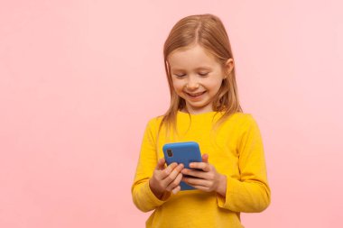 Akıllı telefonu faiziyle ve gülümsemesiyle kullanan mutlu küçük kızın portresi, cep telefonuyla oyun oynayan, eğlenen, basit uygulamalarla tatmin olan. Stüdyo çekimi pembe arkaplanda izole edildi