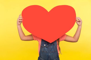 Congratulation on Valentine's day. Portrait of anonymous little girl hiding behind big red heart, holding symbol of love and affection, large greeting card. studio shot isolated on yellow background clipart