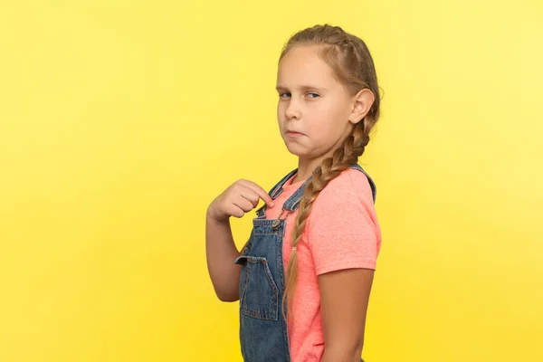 This is me! Portrait of egoistic self-confident little girl in denim overall pointing herself and looking with arrogance, proud of achievement success. indoor studio shot isolated on yellow background