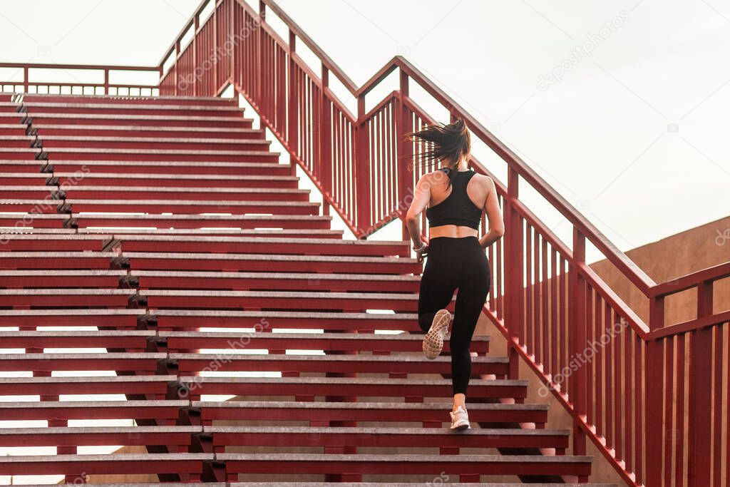 Back view of runner athlete, fit brunette girl in black sportswear, tight pants and top, running up stairs doing cardio training, jogging workouts for weight loss. Health care, sport activity outdoor