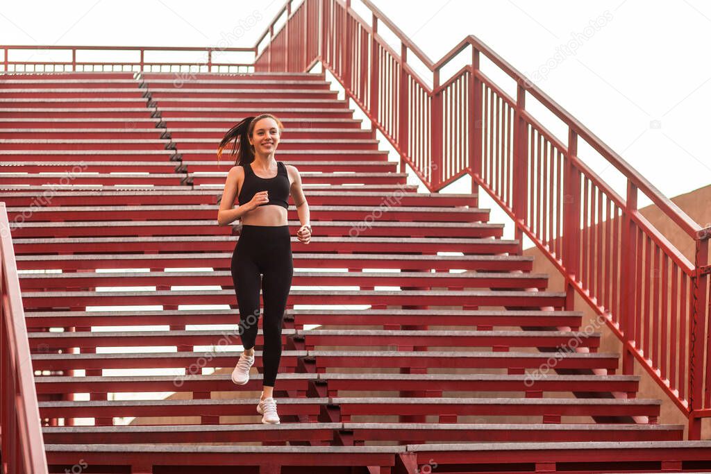 Runner athlete, fit happy girl in black sportswear, tight pants and top, running down stairs doing cardio training, jogging workouts for weight loss. Health care concept, sport activity outdoor