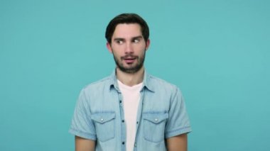 I'm crazy! Bearded guy in jeans shirt with awkward dumb grimace showing stupid gesture, having insane idea, being out of mind, making cuckoo silly face. indoor studio shot isolated on blue background