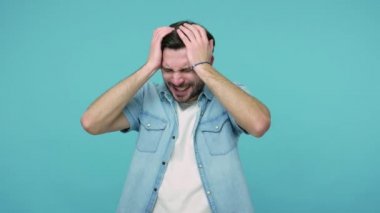 Kotlu tişörtlü, aşırı yorgun sakallı adam ellerini başının üstüne koyuyor, şiddetli baş ağrısı, şiddetli migren ağrısı, ateş ve grip belirtileri gösteriyor. Stüdyo çekimi mavi arkaplanda izole edildi