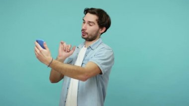 Kot pantolonlu olumlu sakallı adam cep telefonuyla arkadaşlarıyla konuşuyor, bir şeyler anlatıyor, online mobil iletişim kuruyor. Mavi arka planda kapalı stüdyo çekimleri