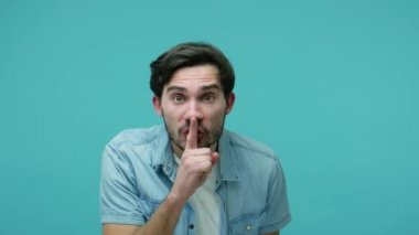 Shh, sessiz ol! Kotlu tişörtlü sakallı adam parmağını dudaklarına koyuyor, sessiz jest yapıyor, üzgün üzgün suratıyla bakıyor, sessizlik istiyor. Mavi arka planda kapalı stüdyo çekimleri