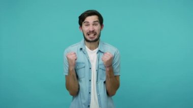 Ben şampiyonum! Kot pantolonlu, aşırı mutlu sakallı çılgın adam yumruk sallıyor ve mutluluktan bağırıyor, evet başarısı, zaferi kutluyor. Mavi arka planda kapalı stüdyo çekimleri